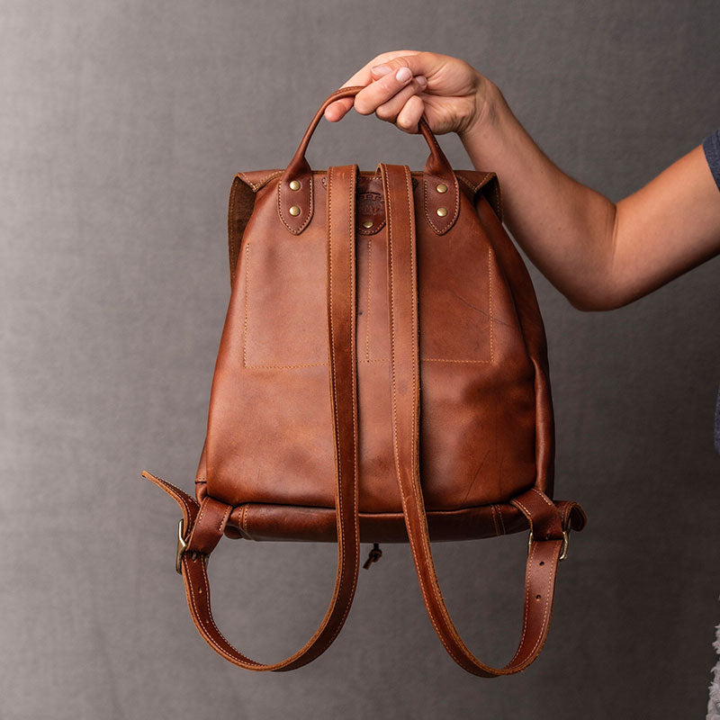 Hand made full grain leather backpack with drawstring slap closure. It features a main compartment with an interior pocket, two outer pockets, and a side pocket. Available in black or brown leather.